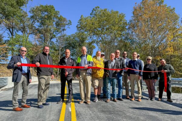 A group of people stands on a newly paved road for a ribbon-cutting ceremony, holding a large red ribbon. Trees and a clear sky are in the background.