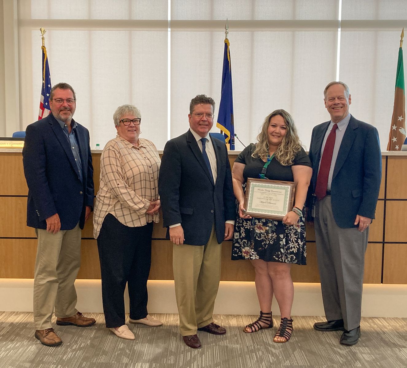 Five people standing in a room, one holding a framed certificate, smiling for a group photo. Flags are visible in the background.