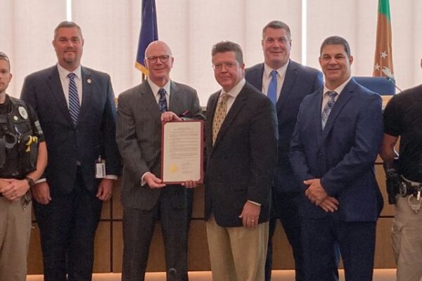 A group of people, some in suits and others in uniforms, stand in a room. One person is holding a framed document. Flags and a wooden bench are visible in the background.