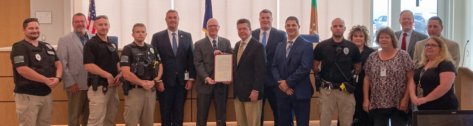 A group of people, some in suits and others in uniforms, stand in a room. One person is holding a framed document. Flags and a wooden bench are visible in the background.