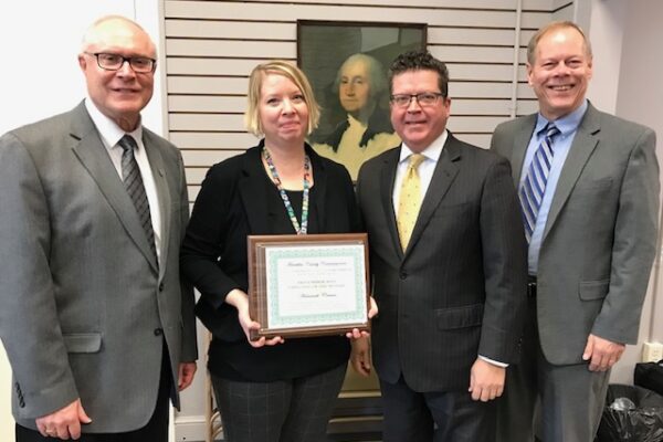 Four people stand together in a room; one holds a framed certificate. A portrait of a historical figure hangs in the background.