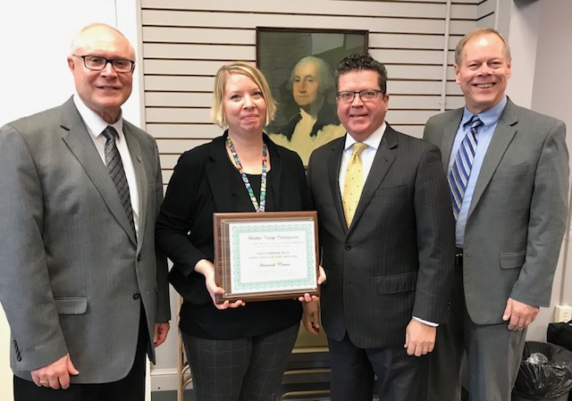 Four people stand together in a room; one holds a framed certificate. A portrait of a historical figure hangs in the background.