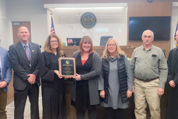 A group of seven people, including two judges, stand in a courtroom. One person holds a plaque. An American flag and state seal are in the background.