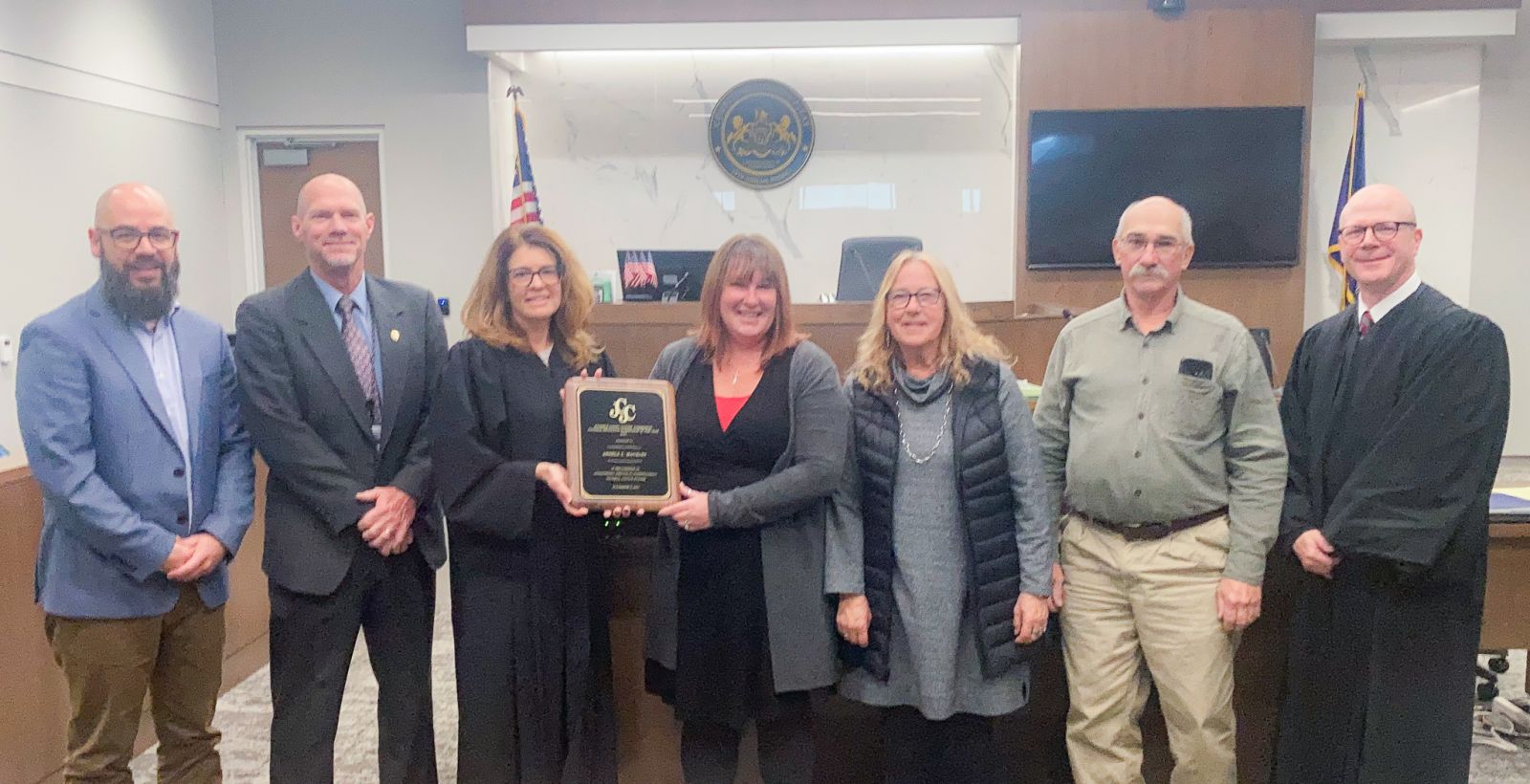 A group of seven people, including two judges, stand in a courtroom. One person holds a plaque. An American flag and state seal are in the background.