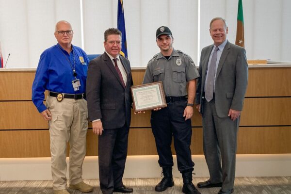 Four men are standing indoors; one holds a framed certificate. Two are in suits, one in a uniform, and one in a blue shirt with a badge and ID. Flags and a wooden panel are in the background.