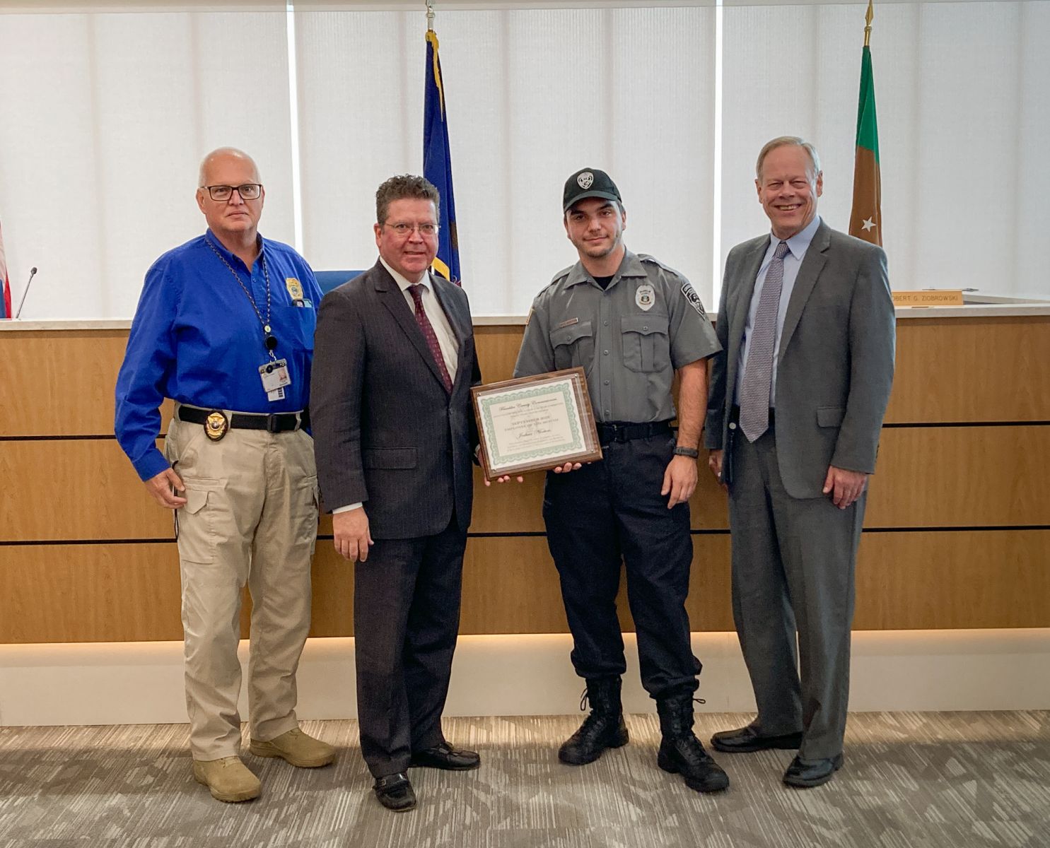 Four men are standing indoors; one holds a framed certificate. Two are in suits, one in a uniform, and one in a blue shirt with a badge and ID. Flags and a wooden panel are in the background.