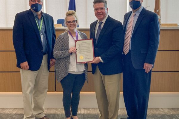 Four people stand together in an office, two men wearing masks, one holding a framed certificate.