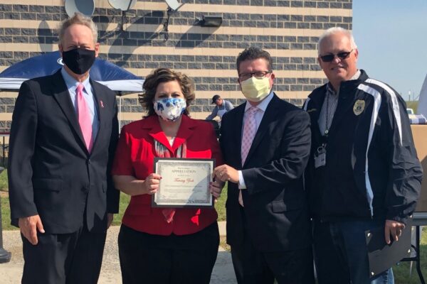 Four people standing outdoors, three in suits, one in a jacket. The woman holds a certificate of appreciation. Three wear face masks, one is maskless. A brick building and umbrellas are behind them.