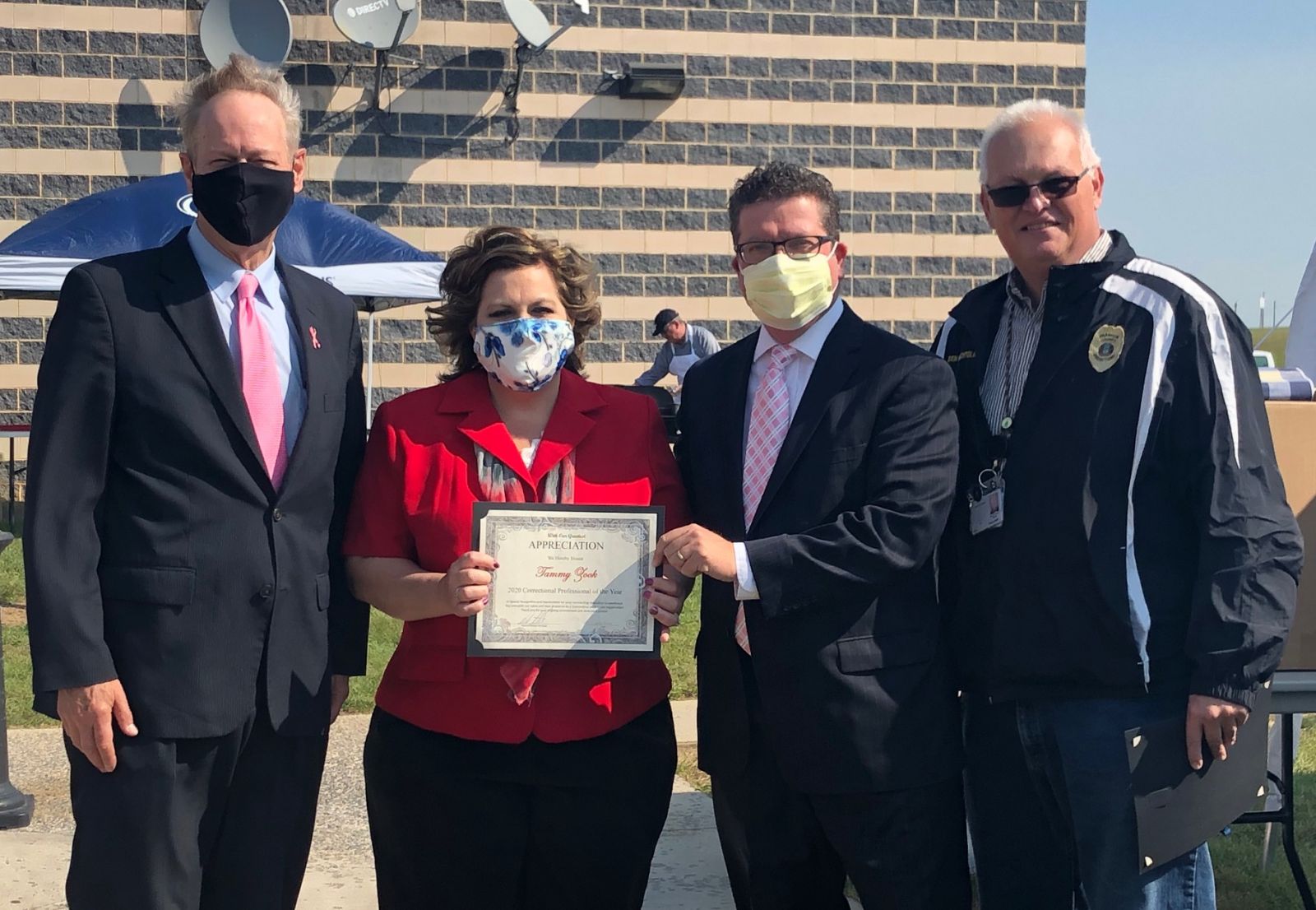 Four people standing outdoors, three in suits, one in a jacket. The woman holds a certificate of appreciation. Three wear face masks, one is maskless. A brick building and umbrellas are behind them.