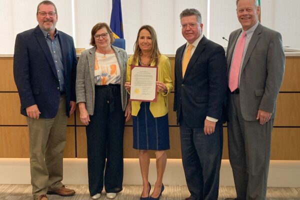 Five people standing together indoors, one woman in the center holding a certificate, all dressed in business attire.