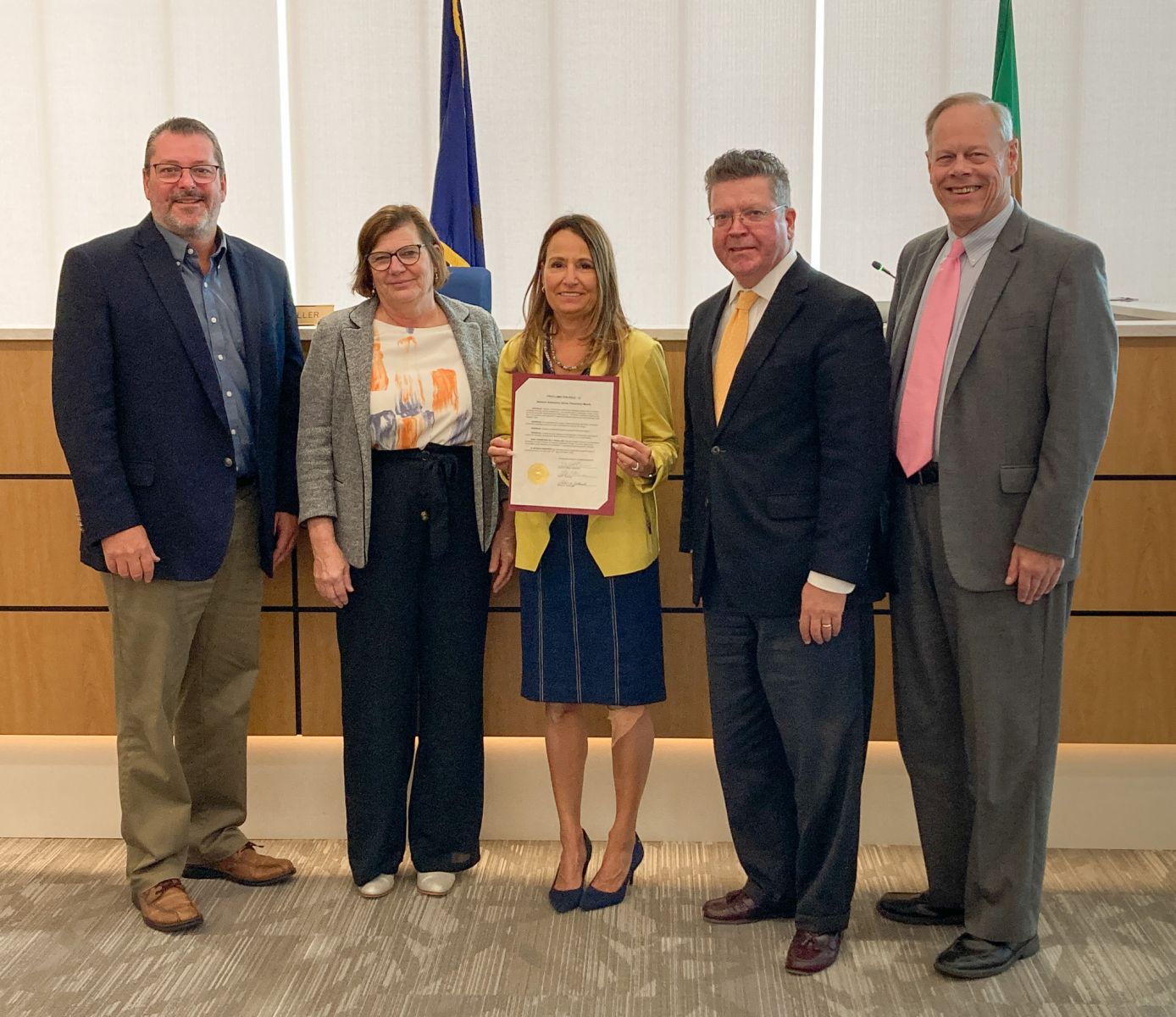 Five people standing together indoors, one woman in the center holding a certificate, all dressed in business attire.