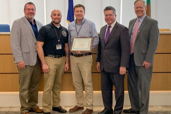 Five men stand in a room, with one holding a framed certificate. They are dressed in business and casual attire. Flags and a wooden desk are visible in the background.