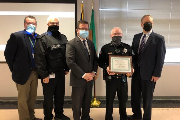 Five men, including two in sheriff uniforms and three in suits, stand in an office. One officer holds a framed certificate. They are all wearing face masks.