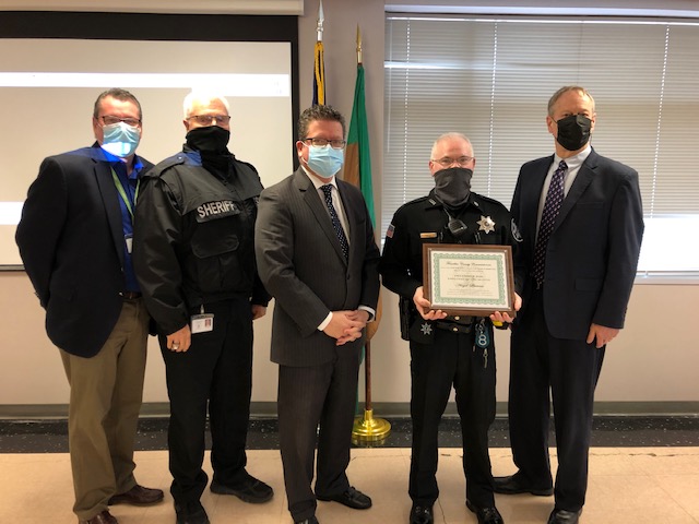 Five men, including two in sheriff uniforms and three in suits, stand in an office. One officer holds a framed certificate. They are all wearing face masks.