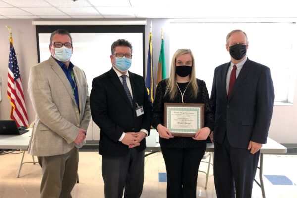 Four people pose indoors, one holding a framed certificate. All are wearing formal attire and masks. Two flags are in the background.