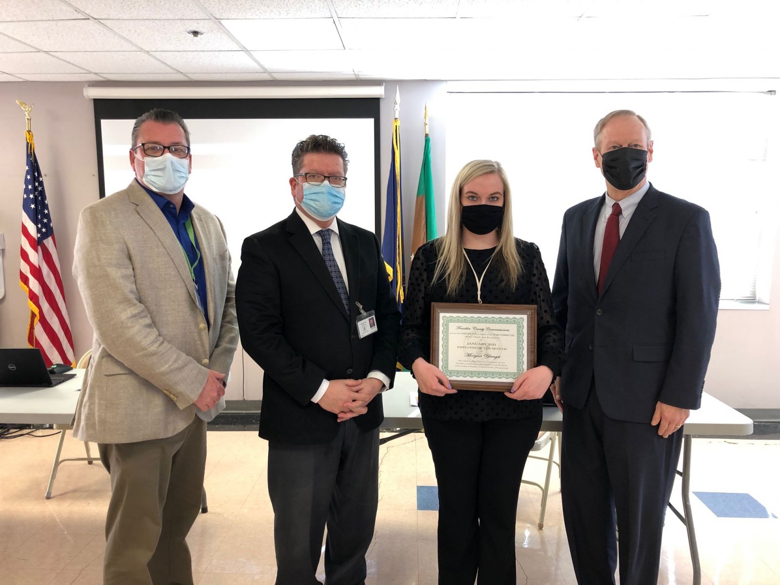 Four people pose indoors, one holding a framed certificate. All are wearing formal attire and masks. Two flags are in the background.