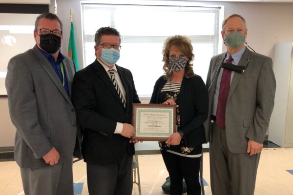 Four people standing together; two holding a framed certificate. Three men and one woman, all wearing masks, with a projection screen and a flag visible in the background.