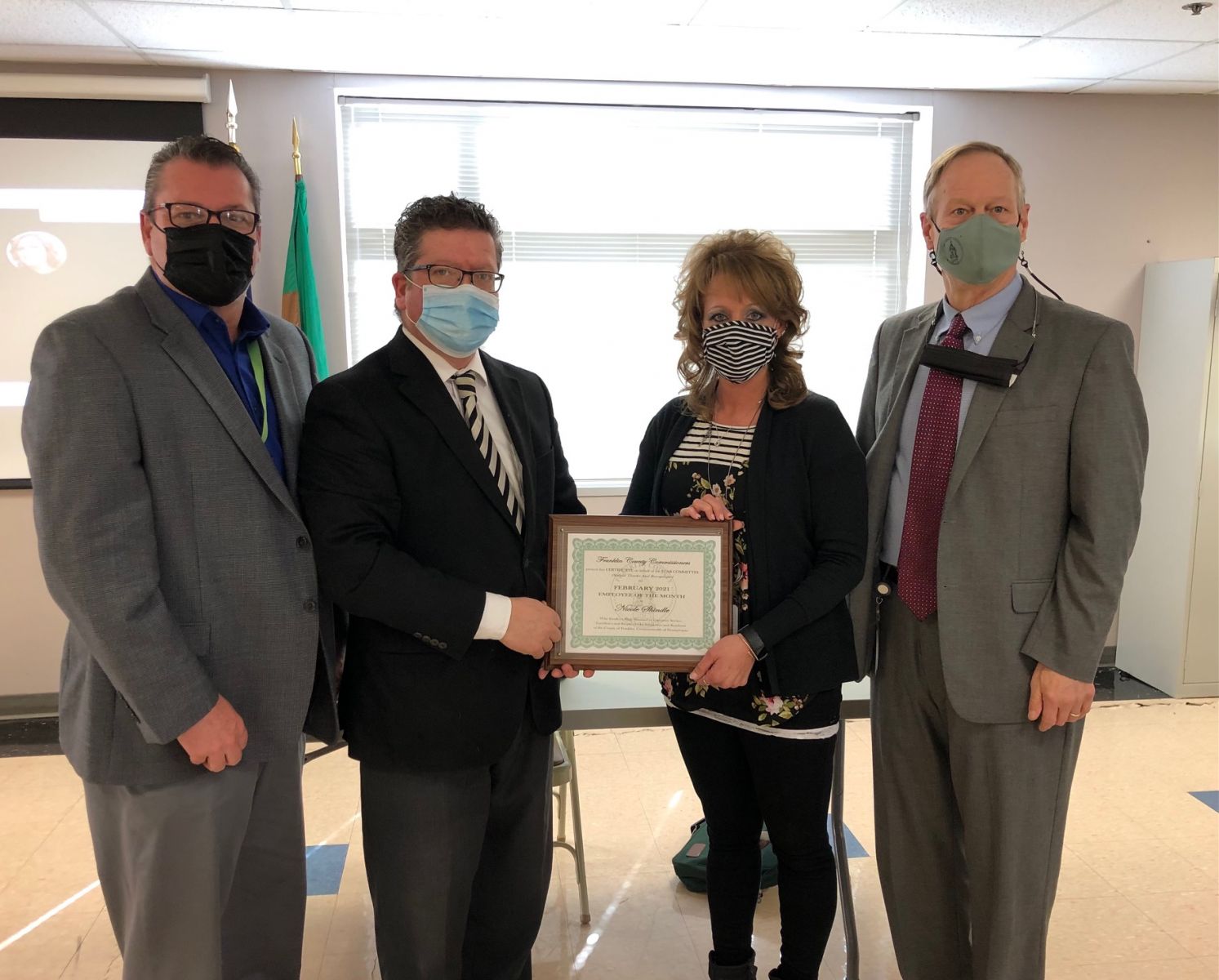 Four people standing together; two holding a framed certificate. Three men and one woman, all wearing masks, with a projection screen and a flag visible in the background.