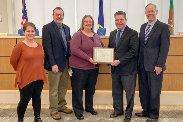 Five people standing indoors, one holding a framed certificate. A U.S. flag and other flags are displayed in the background.