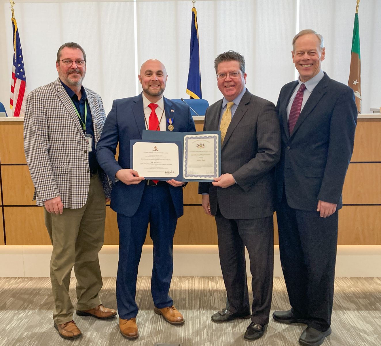 Four men stand side by side in a formal setting, with two holding framed certificates. Flags are visible in the background.