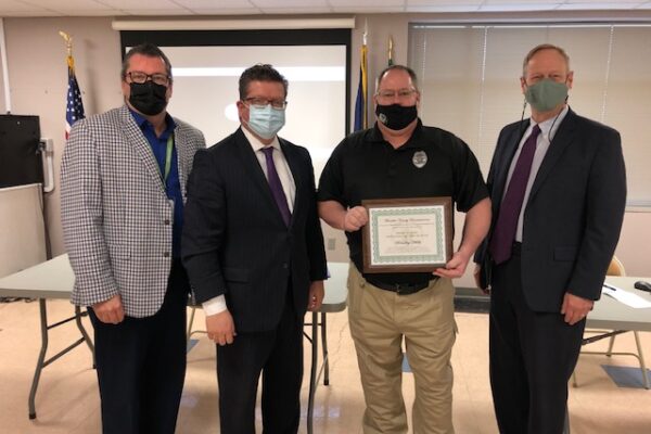 Four men in business attire stand indoors, three wearing masks. One man holds a certificate. A projection screen and flags are in the background.