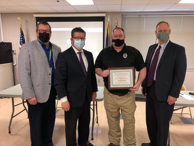 Four men in business attire stand indoors, three wearing masks. One man holds a certificate. A projection screen and flags are in the background.