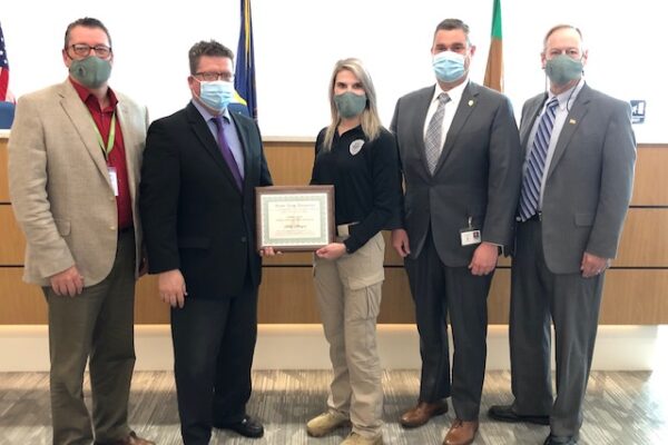 Five people wearing masks stand in a line. The person in the center holds a framed certificate. Flags are visible in the background.