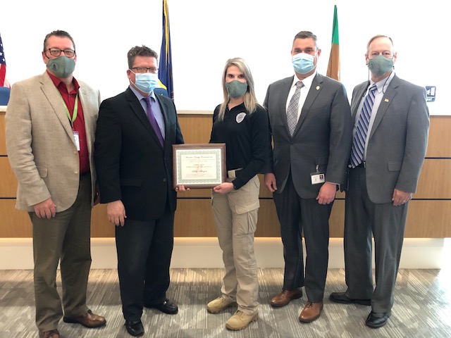 Five people wearing masks stand in a line. The person in the center holds a framed certificate. Flags are visible in the background.