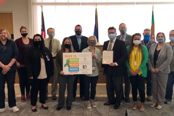 A group of people, some holding signs and a document, pose indoors in a room with flags. They're wearing masks and standing in two rows.