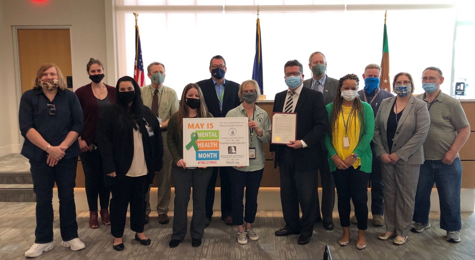 A group of people, some holding signs and a document, pose indoors in a room with flags. They're wearing masks and standing in two rows.