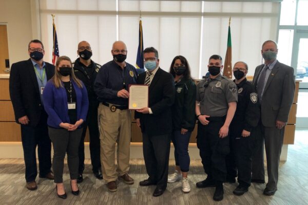 A group of nine people, including law enforcement and office personnel, stand together in a room. One person in the center holds a document. Some people are wearing masks.