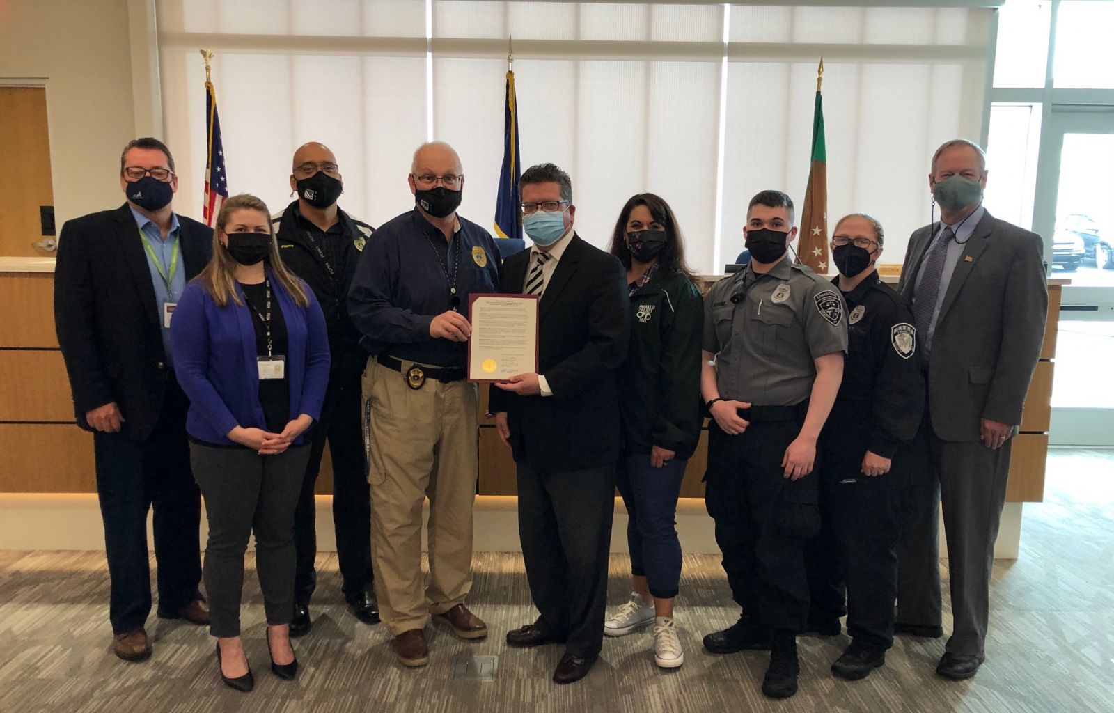 A group of nine people, including law enforcement and office personnel, stand together in a room. One person in the center holds a document. Some people are wearing masks.