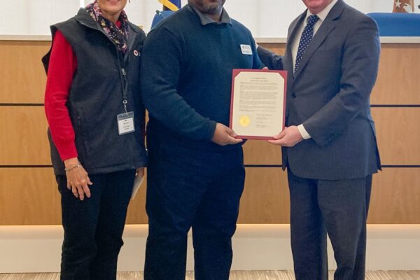 Three people stand indoors. The person in the center holds a certificate. They are in front of a wooden panel and near a flag.