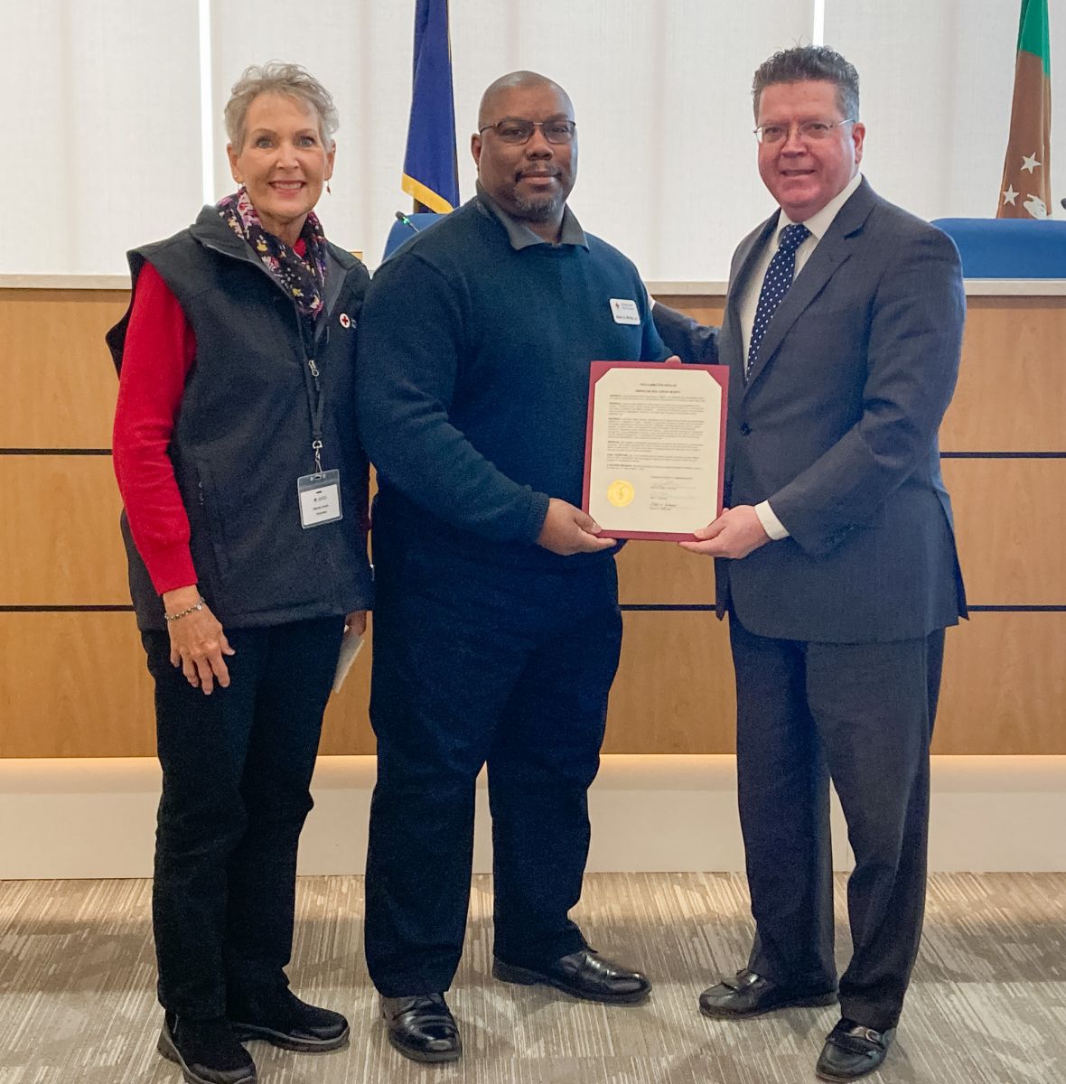 Three people stand indoors. The person in the center holds a certificate. They are in front of a wooden panel and near a flag.