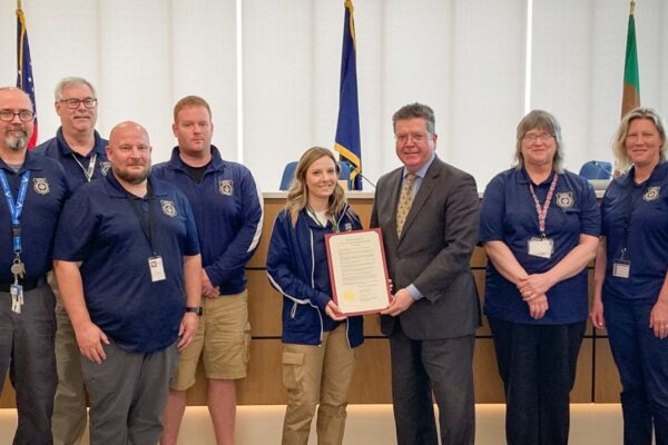 Group of people standing together, some in blue uniforms, with one person holding a framed document. Flags and a wooden desk in the background.