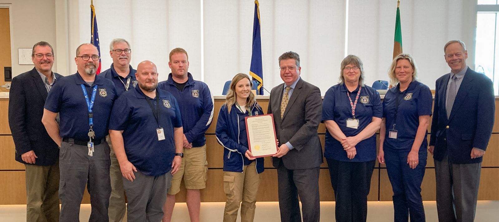 Group of people standing together, some in blue uniforms, with one person holding a framed document. Flags and a wooden desk in the background.