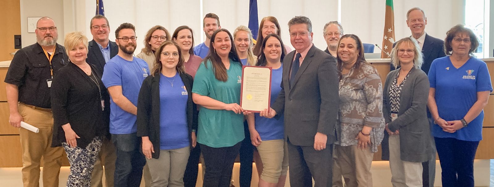 A group of people stand together in a room, with one person holding a certificate. They appear to be in a formal setting.