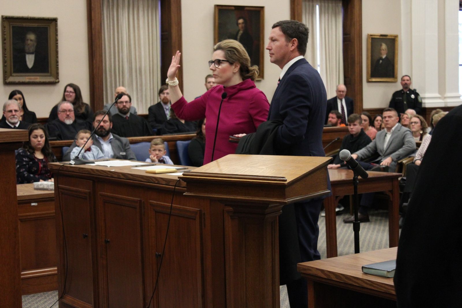 A woman in a pink outfit raises her hand while standing beside a man in a courtroom. Audience members sit in the background.