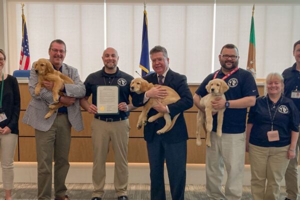 A group of eight people, some holding puppies, stand in a room with flags and a certificate.