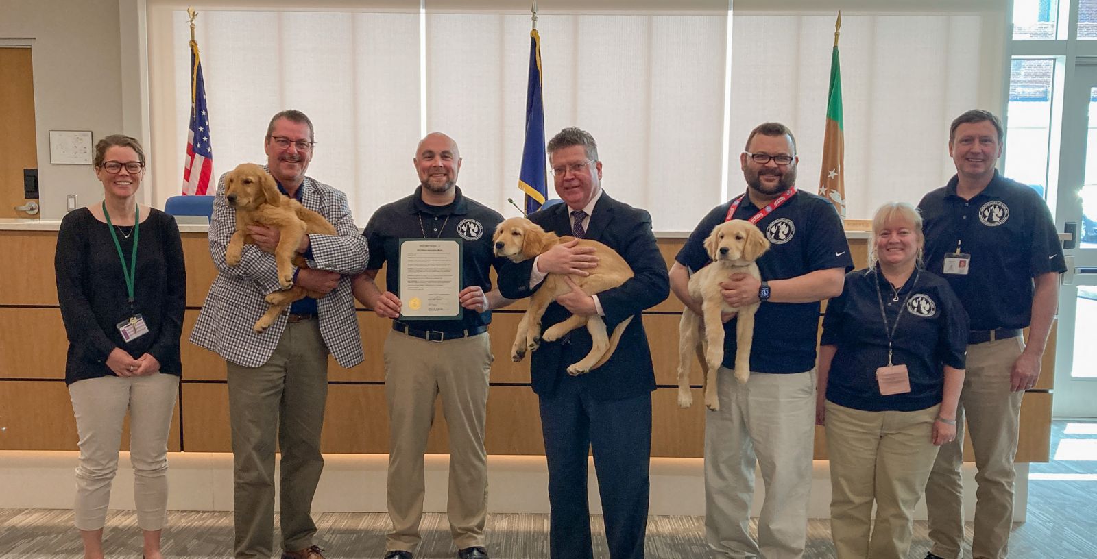 A group of eight people, some holding puppies, stand in a room with flags and a certificate.