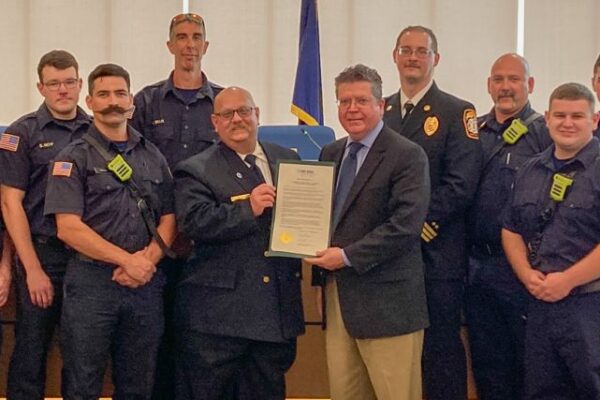 A group of firefighters and officials in uniform pose indoors with a certificate.