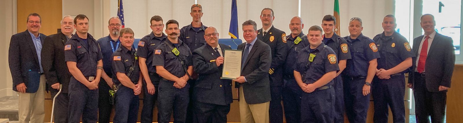 A group of firefighters and officials in uniform pose indoors with a certificate.