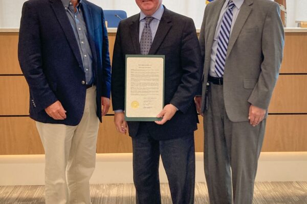 Three men in suits stand together; the man in the center holds a framed document.