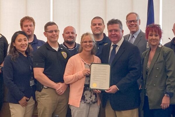 A diverse group of people standing in a room, with some holding a document. Flags are visible in the background.