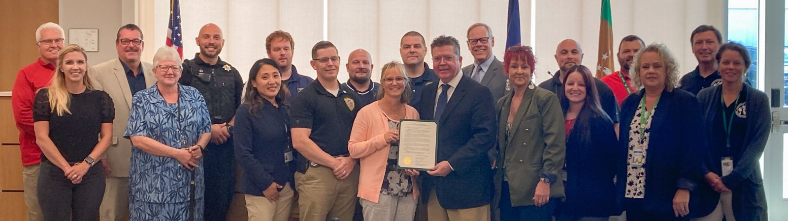 A diverse group of people standing in a room, with some holding a document. Flags are visible in the background.