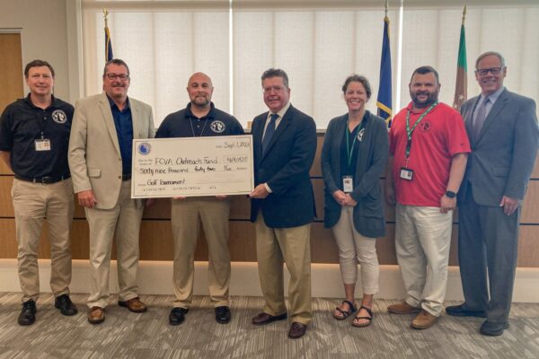 A group of eight people stands in a room. The central person is holding a large ceremonial check, with text indicating a donation to an 