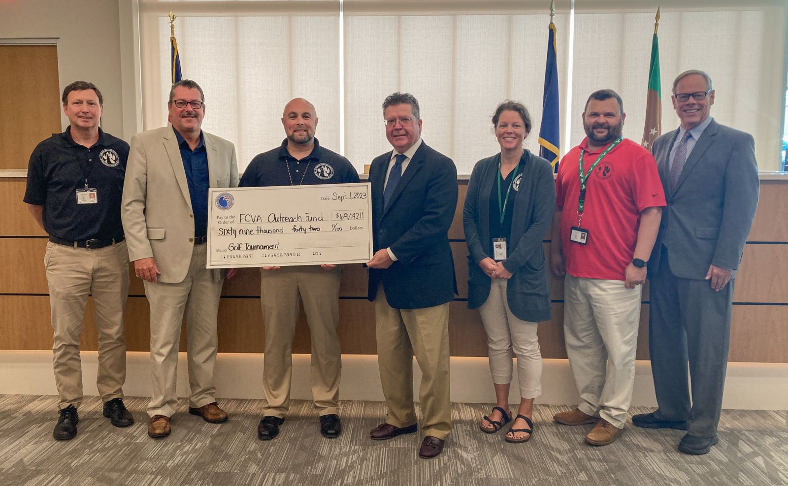 A group of eight people stands in a room. The central person is holding a large ceremonial check, with text indicating a donation to an 