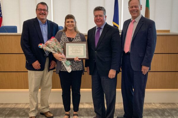 FourPictured (from left): Franklin County Commissioner John Flanner, September Employee of the Month Christina Daywalt, Commissioner Chairman Dave Keller, and Commissioner Bob Ziobrowski people standing together in a room. One person is holding a certificate and flowers. There's a podium in the background.