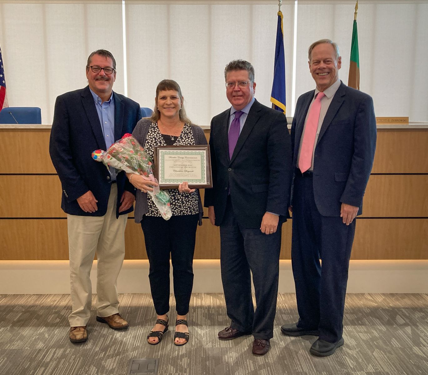 FourPictured (from left): Franklin County Commissioner John Flanner, September Employee of the Month Christina Daywalt, Commissioner Chairman Dave Keller, and Commissioner Bob Ziobrowski people standing together in a room. One person is holding a certificate and flowers. There's a podium in the background.