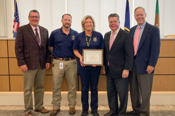 Five people stand in a room; the center person holds a framed certificate. Flags and a wood-paneled wall are in the background.